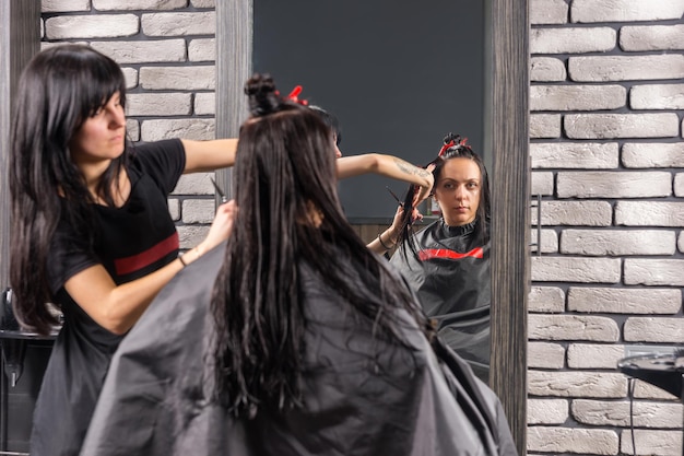 Parrucchiere femminile che taglia e modella i capelli castani con le forbici e il pettine di giovane donna bruna mentre è seduta in poltrona nel salone di bellezza