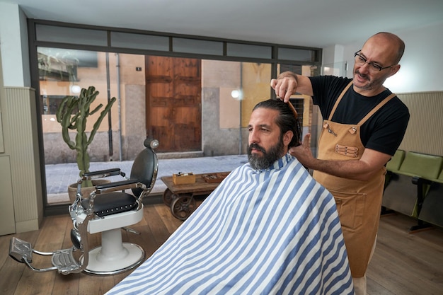 Parrucchiere fiducioso in grembiule che pettina i capelli lunghi di hipster maschio seduto sulla sedia nel negozio di barbiere professionista