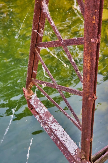 Parte del ponte strutturata e arrugginita con sottili travi metalliche incrociate sull'acqua del fiume con ragnatele