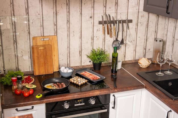 Parte di una cucina contemporanea con lavello e fornello elettrico con pancetta e uova fritte circondate da stoviglie e prodotti alimentari