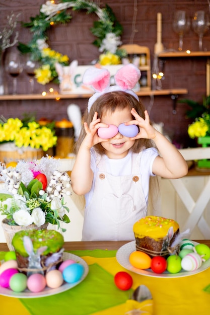Pasqua una bambina gioca con le uova di Pasqua coprendosi il viso con loro nelle orecchie di una lepre e sorride a casa al tavolo festivo a casa in cucina