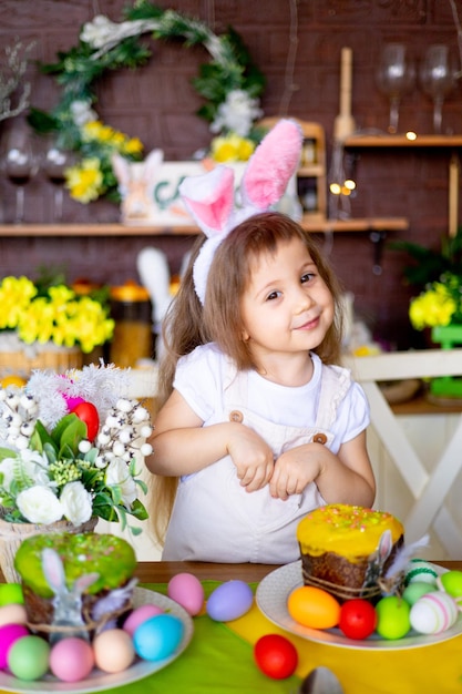 Pasqua una bambina ha piegato le mani come un coniglietto nelle orecchie di un coniglio e sorride a casa a una tavola festiva con uova di Pasqua e una torta a casa in cucina