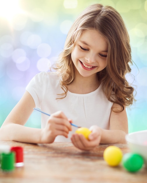 Pasqua, vacanze e concetto di bambino - ragazza felice con pennello che colora le uova di Pasqua su sfondo di luci