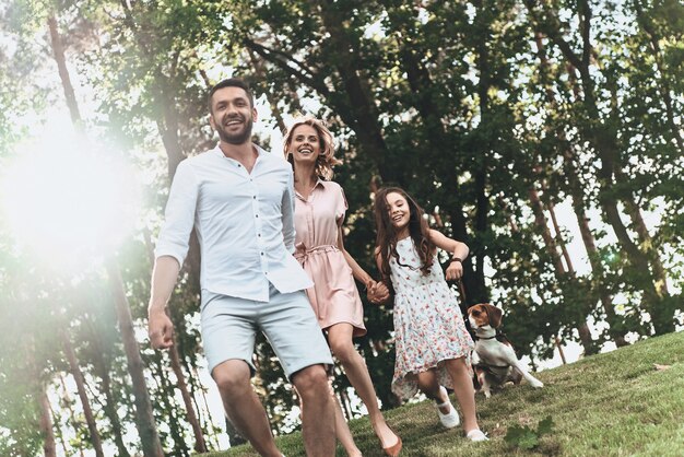 Passare del tempo con la famiglia. Felice giovane famiglia di tre persone con cane che si tiene per mano e sorride mentre corre nel parco