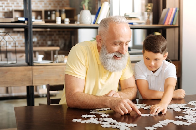 Passatempo rilassante. Felice l'uomo anziano seduto al tavolo accanto al suo nipotino e cercando di fare un grande puzzle con lui