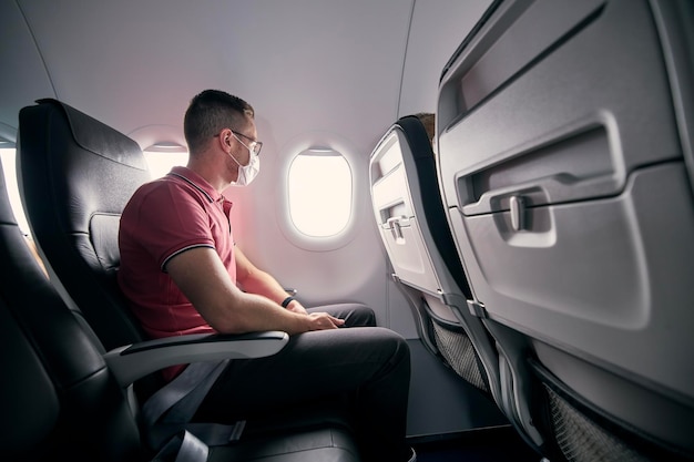 Passeggero con maschera protettiva nell'aereo che guarda attraverso la finestra