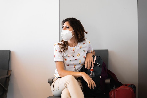 Passeggero femminile seduto all'aeroporto del terminal in attesa con maschera protettiva e bagagli