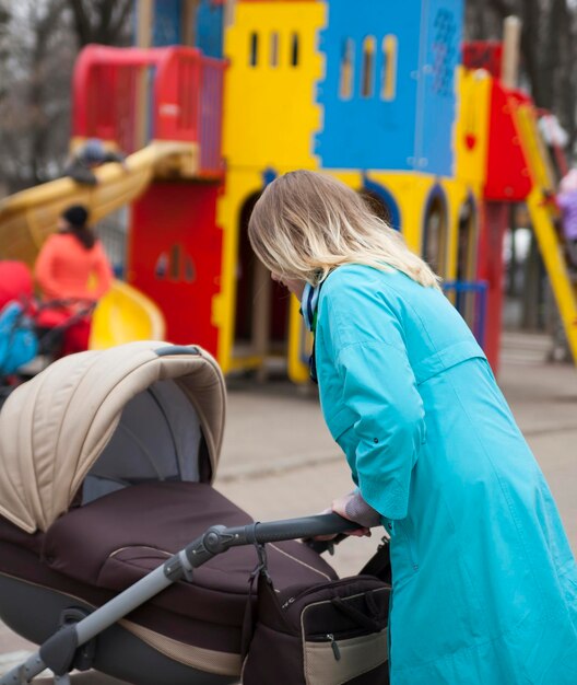 Passeggiata in città con passeggino giovane madre attiva in trench blu che cammina nel parco giochi con il suo bambino in carrozzina