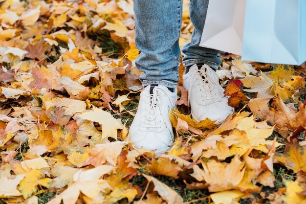 Passeggiata nel parco autunnale Colpo ritagliato di uomo in scarpe da ginnastica in piedi con borse della spesa su foglie d'acero gialle