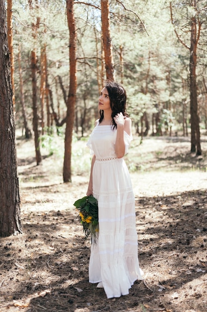 Passeggiata nuziale in pineta. Giorno soleggiato. Sposi nella foresta. Bella sposa e sposo in una passeggiata. Abito da sposa bianco. Bouquet di peonie e ortensie.