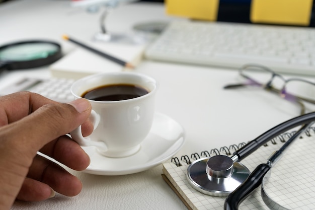 Passi la tenuta della tazza di caffè con gli oggetti business sulla tavola del cuoio bianco.