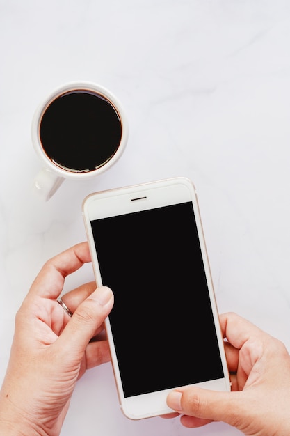 Passi lo smartphone della tenuta con la tazza di caffè nero su fondo di marmo bianco