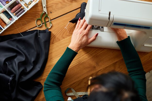 Passo dopo passo, una donna di 50 anni cuce vestiti sulla macchina da cucire, sarto maturo che lavora con il cucito in atelier, industria tessile, hobby, spazio di lavoro. Processo di creazione fai da te, posto di lavoro di sarta.