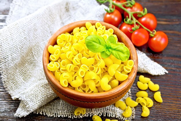 Pasta al gomito con basilico in una ciotola di argilla sul tovagliolo di tela, pomodori sul fondo del bordo di legno scuro