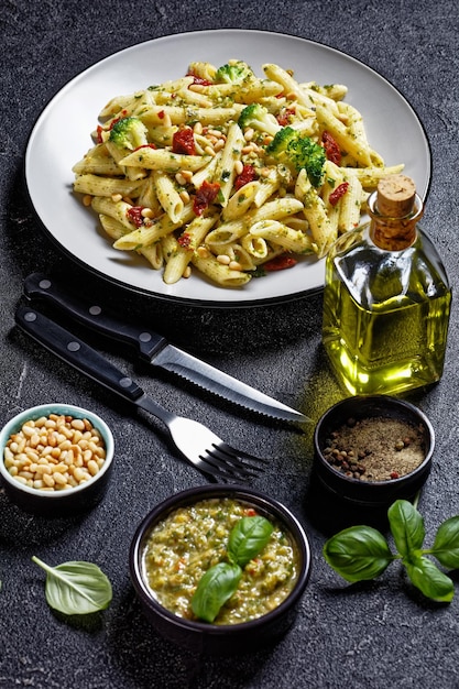 Pasta al pesto di broccoli con pomodori secchi e pinoli su un piatto e ingredienti su un tavolo di cemento, vista verticale dall'alto