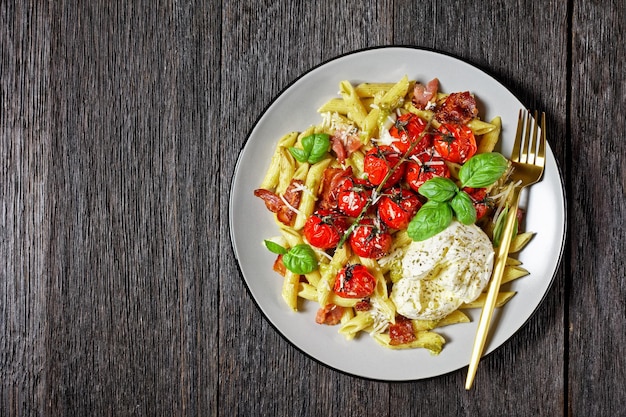 Pasta caprese italiana al forno di penne, pancetta, pomodorini e mozzarella, parmigiano grattugiato, basilico fresco servito su un piatto su un tavolo di legno scuro, vista dall'alto, primo piano, spazio per la copia