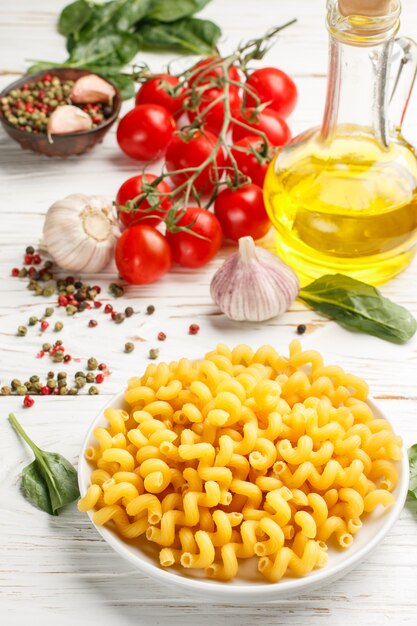 Pasta cruda e ingredienti per cucinare la cena - spinaci, pomodoro, aglio, spezie e olio d'oliva sulla tavola di legno bianca