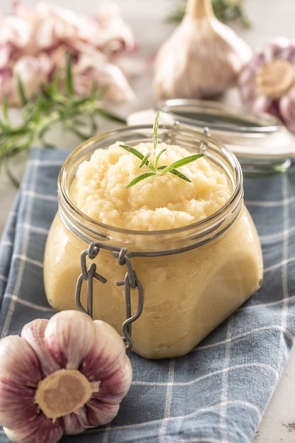 Pasta di aglio aromatica in un barattolo di vetro adagiato su un panno da cucina rustico con bulbi e chiodi di garofano sbucciati e rosmarino.