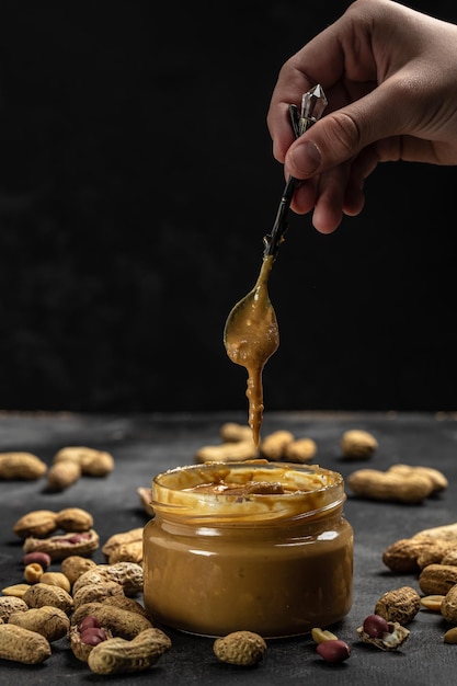 Pasta di arachidi in un barattolo aperto in un cucchiaio vicino a noccioline cremose al burro di arachidi sparse su uno sfondo scuro posto per la vista dall'alto del testo