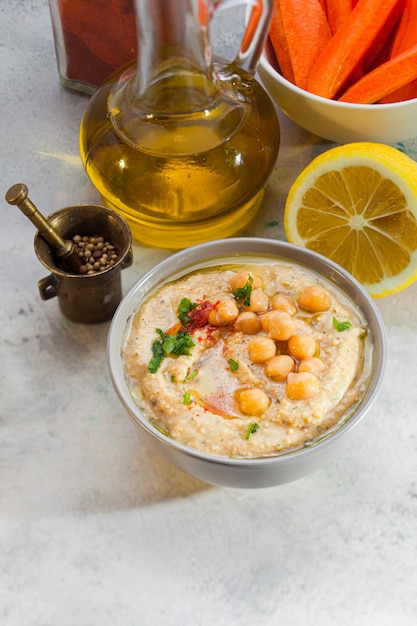 Pasta di hummus in ciotola bianca decorata con olio e verde Mortaio di rame vasetto in vetro con olio d'oliva limone e ciotola con carote sbucciate Spuntino gustoso in stile orientale