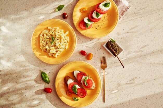 Pasta mediterranea fusilli insalata caprese pomodori mozzarella basilico e pesto sotto le ombre dell'angolo alto Antipasto di pane con insalata caprese su piatti d'arancia Vista dall'alto cibo piatto disteso
