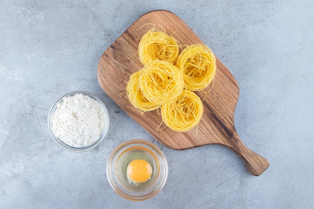 Pasta secca cruda del nido con l'uovo crudo e una ciotola di vetro di farina su una superficie di pietra.