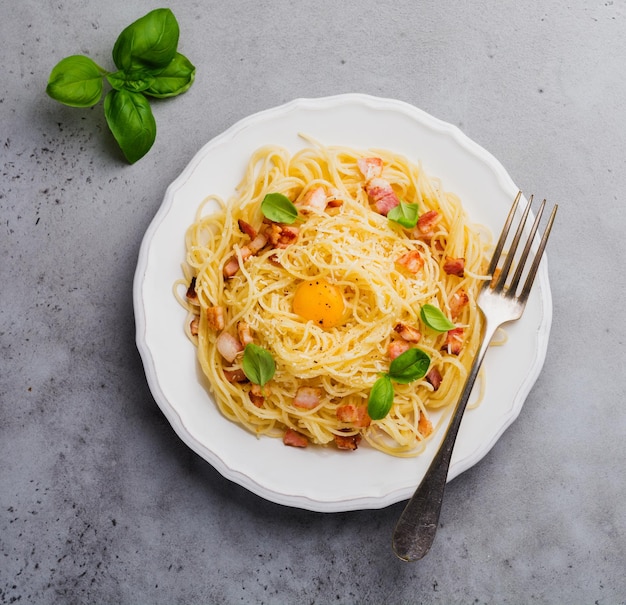 Pasta spaghetti alla carbonara con pancetta, parmigiano, tuorlo d'uovo e foglie di basilico su fondo grigio chiaro. Piatto della tradizione italiana. Vista dall'alto.