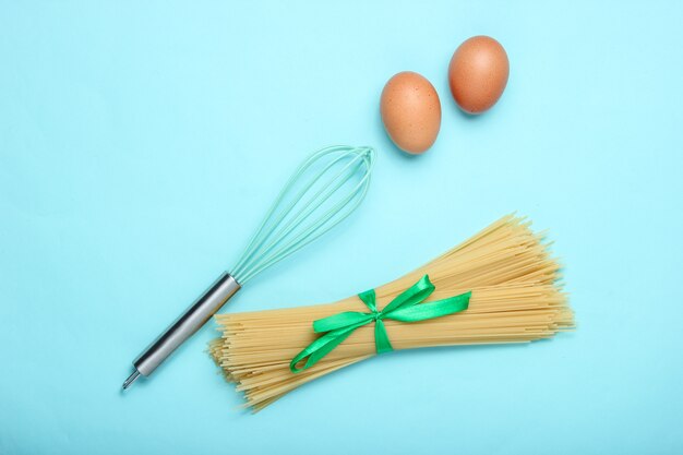 Pasta, uova di gallina e frusta sul blu. Vista dall'alto, stile piatto