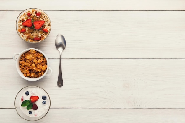 Pasti sani per la colazione sullo spazio della copia del tavolo di legno. Cornflakes, muesli e yogurt con frutti di bosco e cucchiaio, vista dall'alto