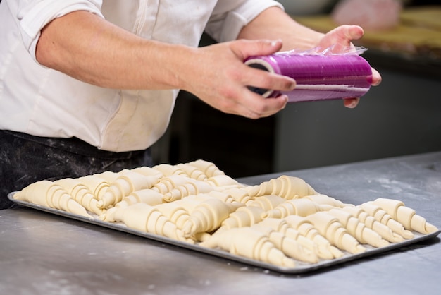 Pasticcere che sigilla con pellicola di plastica un vassoio di pasta per croissant.