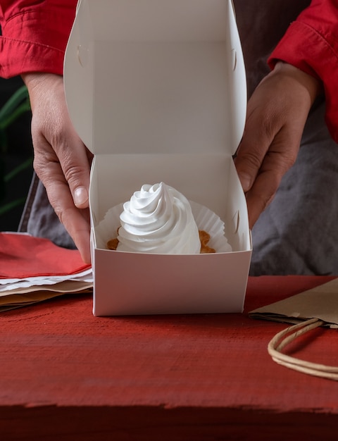 Pasticcere che tiene la scatola di carta bianca con torta bianca vicino al tavolo rosso.