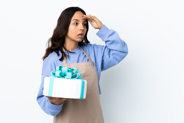 Pasticcere che tiene una grande torta sopra bianco isolato con l'espressione di sorpresa mentre guarda il lato