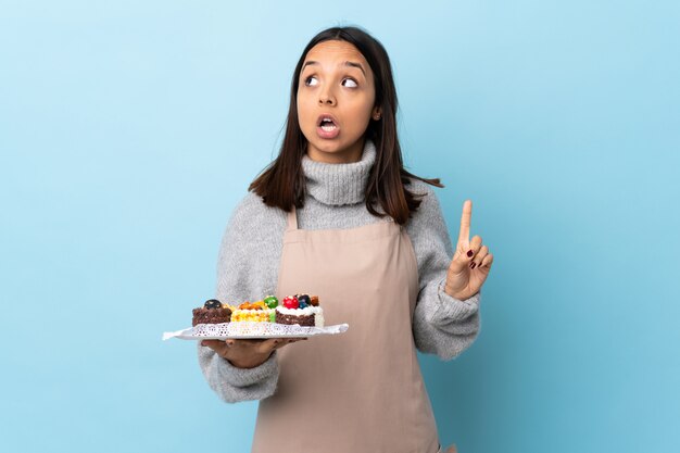 Pasticcere che tiene una grande torta sopra la parete blu isolata che pensa un'idea che indica il dito su