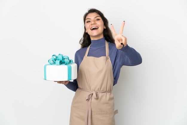 Pasticcere chef russo tenendo una grande torta isolata su sfondo bianco sorridendo e mostrando il segno di vittoria