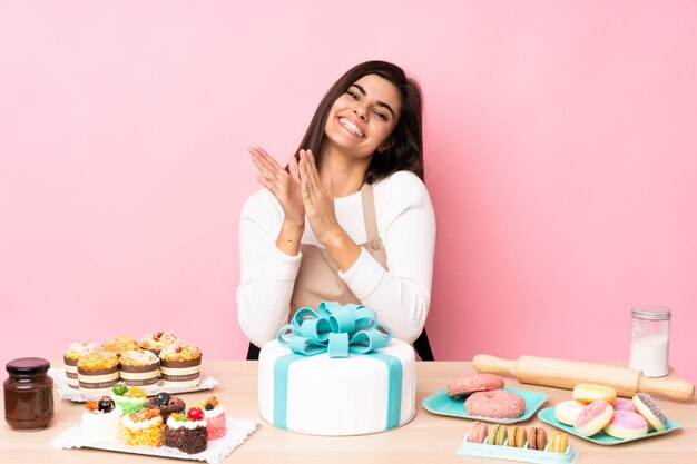 Pasticcere con un grande dolce in una tavola sopra la parete rosa isolata che applaude dopo la presentazione in una conferenza