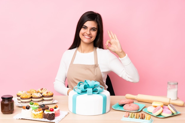 Pasticcere con un grande dolce in una tavola sopra la parete rosa isolata che mostra segno giusto con le dita