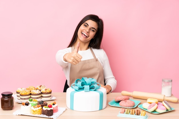 Pasticcere con una grande torta in un tavolo sopra il rosa isolato con il pollice in alto perché è successo qualcosa di buono