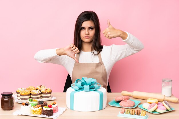 Pasticcere con una grande torta in un tavolo sopra la parete rosa isolata che fa segno buono-cattivo. Indeciso tra sì o no