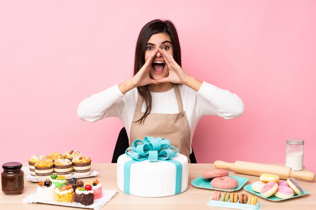 Pasticcere con una grande torta in una tabella sopra la parete rosa isolata che grida e che annuncia qualcosa