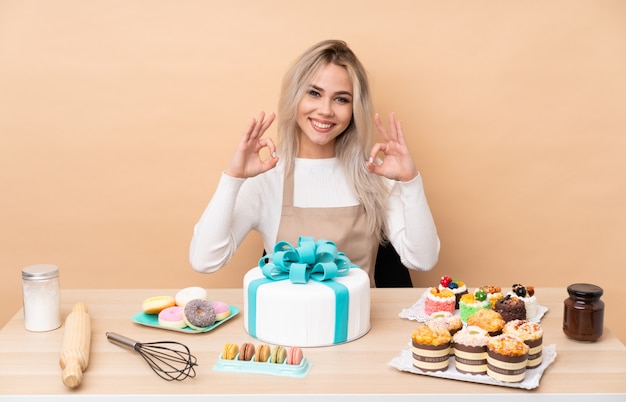 Pasticcere dell'adolescente con una grande torta in una tabella che mostra un segno giusto con le dita