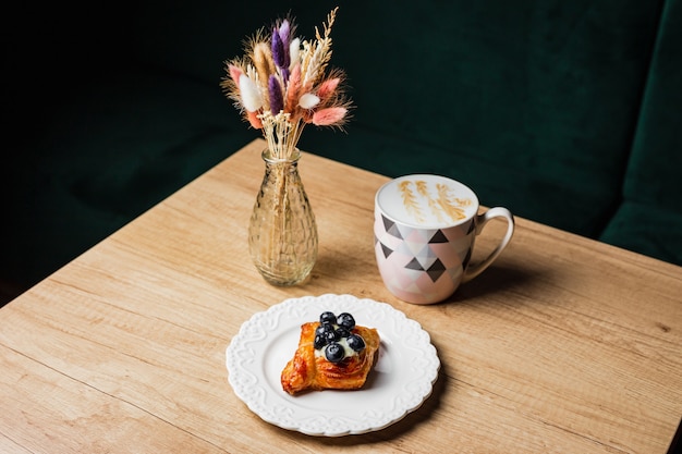 Pasticceria danese con crema pasticcera e palude di mirtilli su un piatto bianco, una tazza di caffè bianco piatto e fiori in un vaso