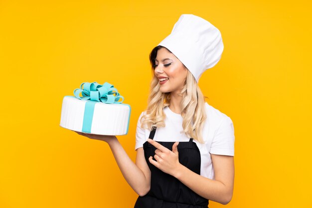 Pasticceria della giovane donna che giudica una grande torta isolata sulla parete gialla che indica dito il lato