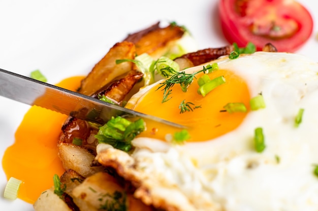 Patate fritte con uovo con tuorlo liquido, erbe aromatiche e pomodori su un piatto bianco.