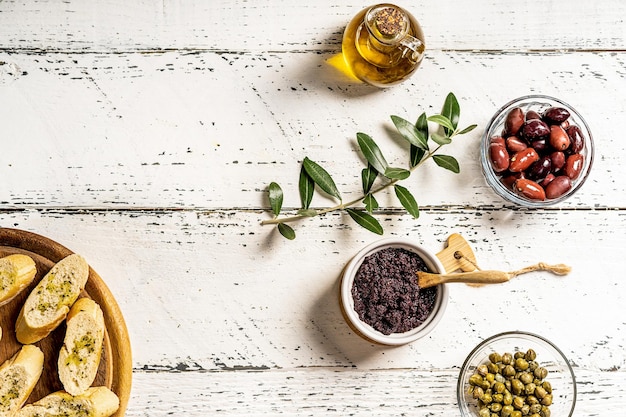 Patè di olive in ciotola, olio d'oliva, olive e capperi su fondo di legno bianco