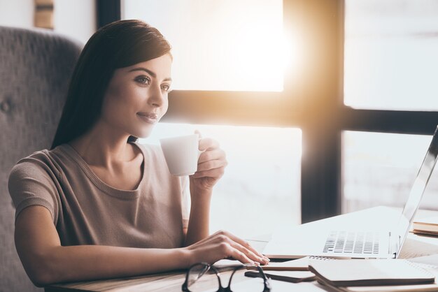 Pausa caffè al lavoro. Giovane bella donna che tiene una tazza di caffè e guarda lontano mentre è seduta su una sedia al suo posto di lavoro