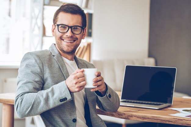 Pausa caffè. Bel giovane con gli occhiali che tiene una tazza di caffè e guarda la telecamera con un sorriso mentre è seduto al suo posto di lavoro