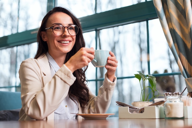 Pausa caffè. Donna allegra piacevole intelligente seduta al tavolo mentre sorride e tiene il caffè