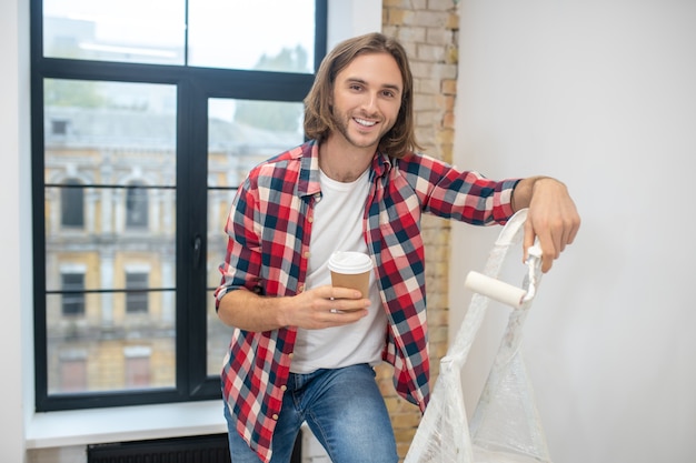 Pausa caffè. Giovane uomo in piedi vicino alla scala a pioli e avendo caffè