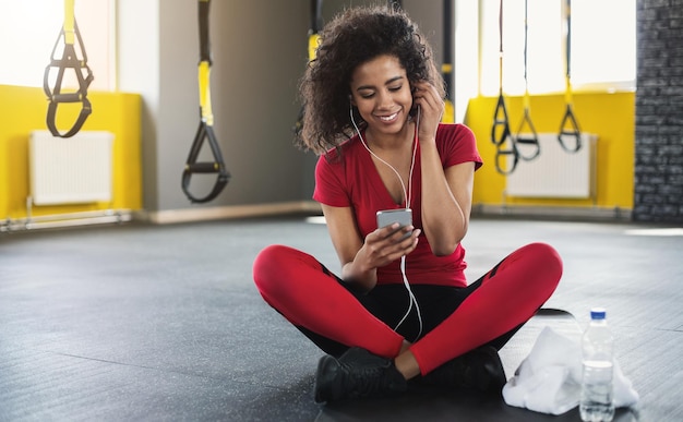 Pausa dopo l'allenamento. Donna sportiva afroamericana che ascolta musica su smartphone in palestra