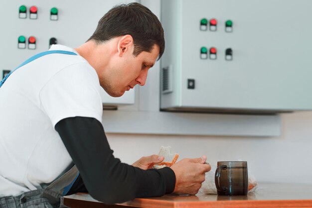 Pausa pranzo Mangiare sul posto di lavoro durante il lavoro L'uomo caucasico in tuta da lavoro si siede al tavolo nella sala di produzione e mangia dal container Il lavoratore pranza nella sala pausa